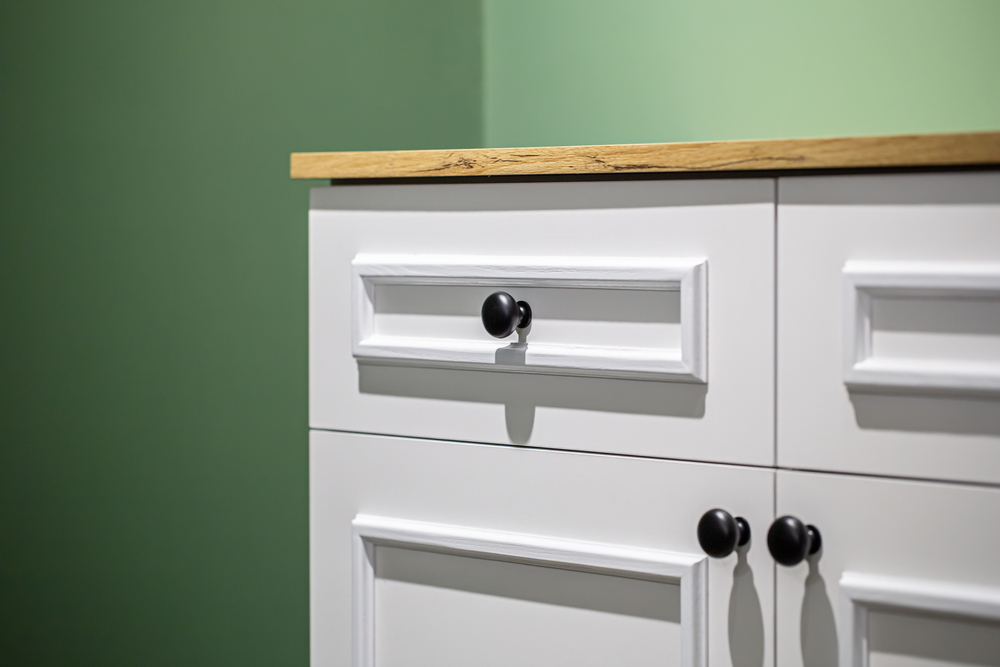 Close-up of a white cabinet with black handles on a green wall background.